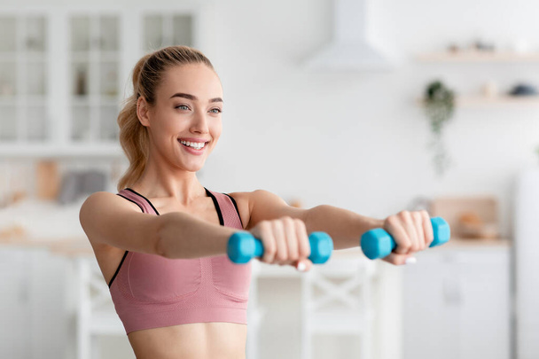 Cheerful glad caucasian pretty young blonde female lifting dumbbells at home, work out indoor and do exercises - Φωτογραφία, εικόνα
