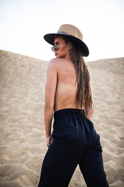 meztelen félmeztelen barna modell fekete nadrágban és kalapban, pózol a homokon a kamera előtt. Közelkép és teljes hosszúságú forgatás. - Fotó, kép
