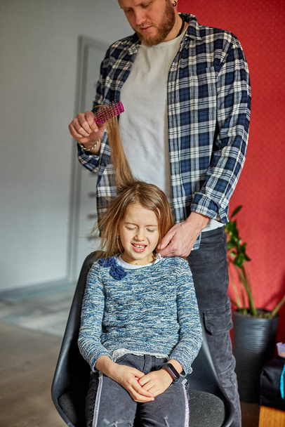 Father combing, brushing his daughter's hair at home, child making faces about hair pulling, angry girl feeling pain, family moments, spending time together. - Photo, Image