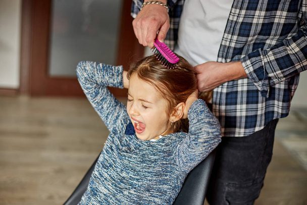 Father combing, brushing his daughter's hair at home, child making faces about hair pulling, angry girl feeling pain, family moments, spending time together. - Foto, Imagen