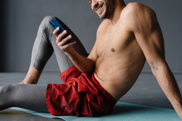 Young shirtless sportsman using mobile phone while working out indoors - Foto, Imagem