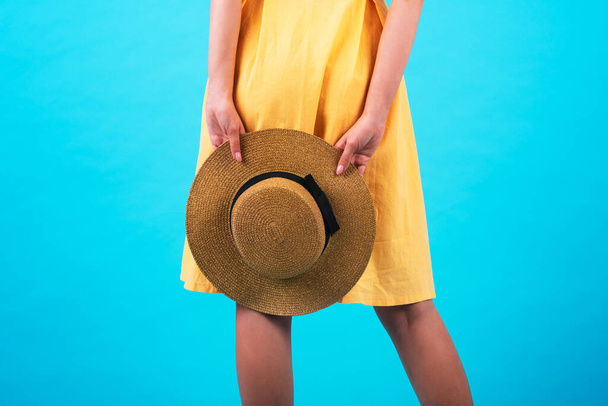 Το κορίτσι κρατάει ένα ψάθινο καπέλο με ένα κίτρινο, φωτεινό φόρεμα σε μπλε φόντο ενός τόνου. Κοντινό πλάνο. Το σύμβολο του ταξιδιού. Ταξιδιωτικό γραφείο. Αντιγραφή χώρου - Φωτογραφία, εικόνα