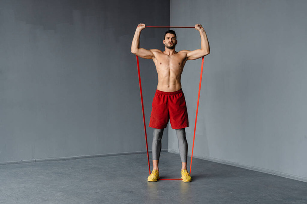 Young shirtless sportsman doing exercise with expander stretch tape indoors - 写真・画像