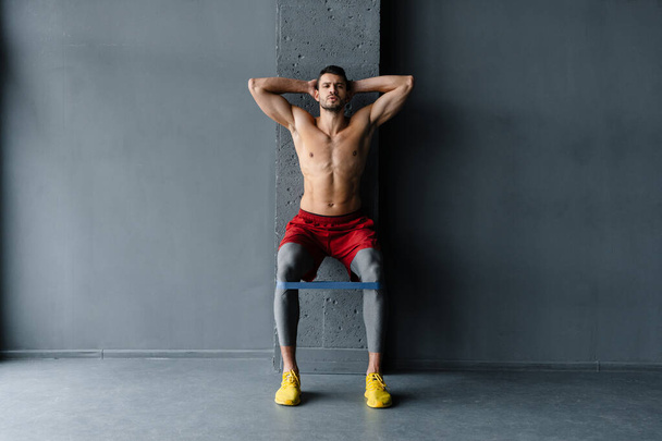 Young shirtless sportsman doing exercise with expander stretch tape indoors - 写真・画像