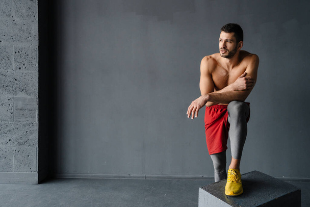 Young muscular shirtless sportsman exercising on a cube indoors resting leaning on a box - Foto, imagen