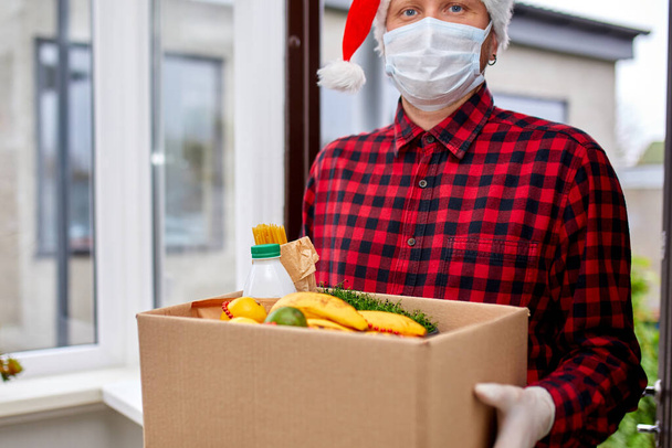 Vrijwilliger in Santa hoed en beschermende masker en handschoenen levering donatie doos thuis in Kerstmis, Vakantie Courier met verpakking doos met voedsel, contactloze levering, - Foto, afbeelding