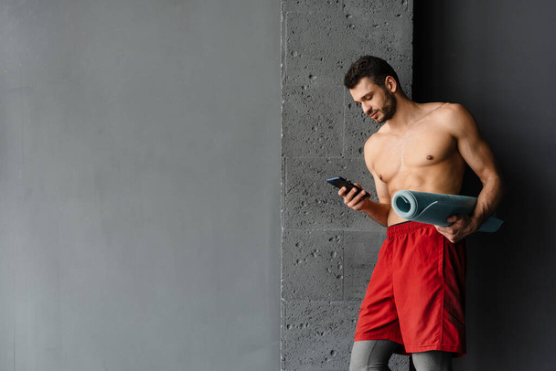 Young shirtless sportsman using cellphone while standing with mat indoors - Foto, immagini
