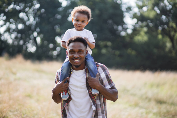 African boy sitting on father shoulders during outdoor walk - Zdjęcie, obraz
