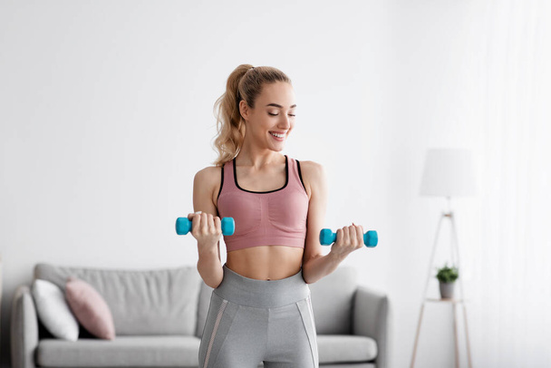 Smiling european beautiful young blonde woman strength exercising with dumbbells at home - Фото, изображение