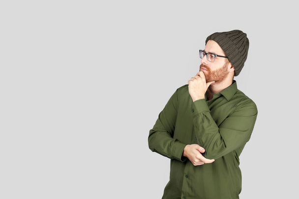Portret młodego Latynosa myślącego, patrzącego w bok, ubranego w zieloną koszulę i wełnianą czapkę, odizolowanego na szarym tle. Przestrzeń kopiowania. - Zdjęcie, obraz