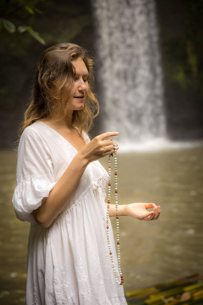 Valkoihoinen nainen pitää lukua rukoilun ja meditaation aikana. Luovuta gyan mudra. Buddhalainen japa-mala. Säikeet jalokiviä helmiä. Uskonnon käsite. Nainen valkoisessa mekossa. Tibumanin vesiputous, Bali - Valokuva, kuva