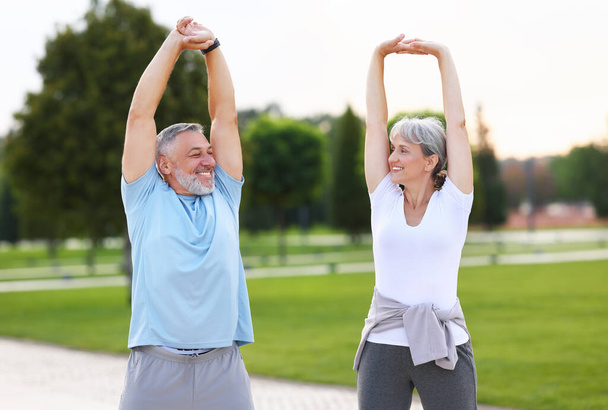 Morning exercises. Happy mature married couple, man and woman in sportswear warming up together doing stretching exercises raised hands up outdoors on park lane in morning. Healthy lifestyle concept - Photo, Image