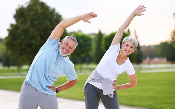 Healthy mind and body. Full length shot of happy smiling mature family man and woman in sportswear stretching arms while warming up together outdoors in park lane on sunny morning. Active lifestyle - Photo, Image