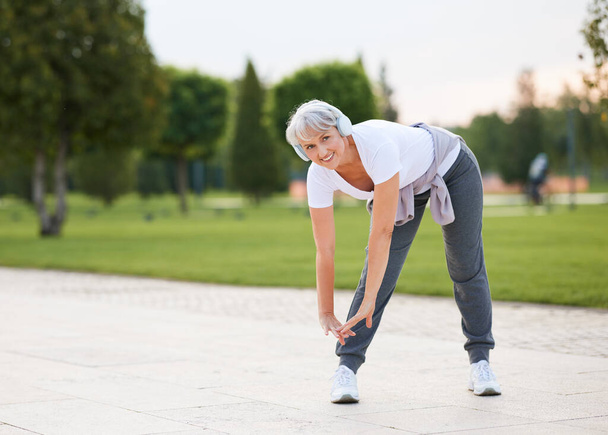 Elderly fit smiling woman in wireless headphones doing stretching exercises before jogging, standing with legs wide apart making forward bends outside in city park alley. Healthy lifestyle concept - Photo, Image