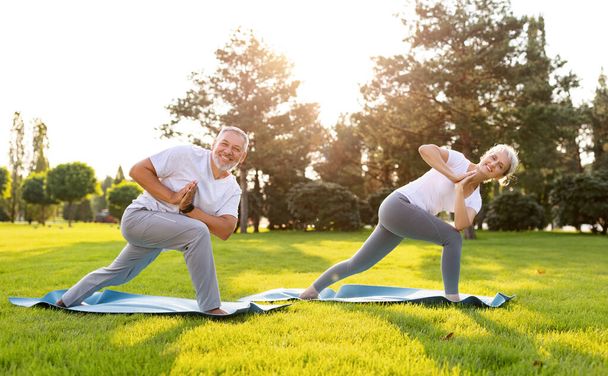 Aktive reife Männer und Frauen machen Partner-Yoga-Übungen im Stehen auf Gummimatten in ausgedehntem Seitenwinkel, praktizieren Asanas im Freien im Park, gesundes, aktives Senioren-Paar trainiert bei Sonnenaufgang - Foto, Bild