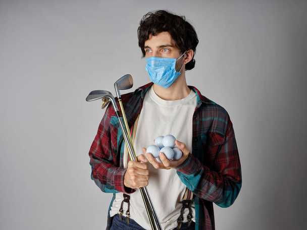 A golfozó alkalmi ruhában és védő kovid-19-es maszkban egy vasat és golflabdát tart a kezében.. - Fotó, kép
