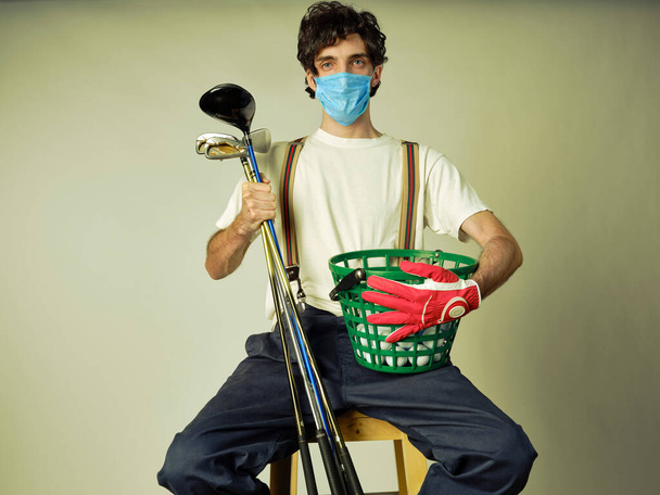 Golf player in casual clothing wearing a protective covid-19 mask, holding some golf irons and a golf drive - Photo, Image