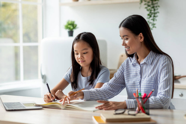 Молода азіатська мати допомагає доньці з домашнім завданням, сидячи за столом в інтер'єрі кухні, вільний простір
 - Фото, зображення