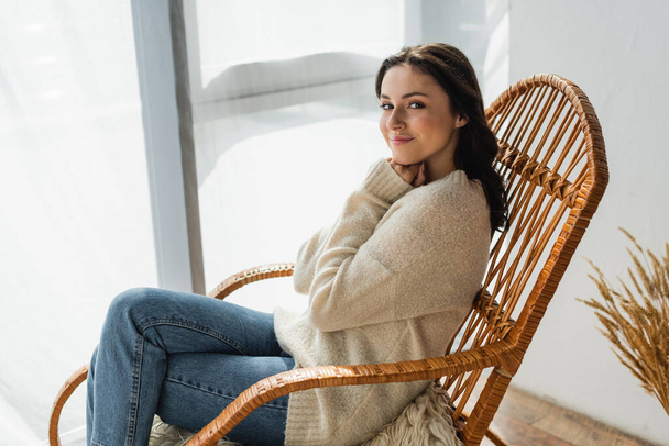 young woman in warm sweater and jeans sitting in wicker chair and smiling at camera - Photo, Image