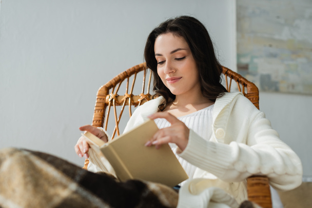 jeune femme souriant tout en lisant le livre dans la chaise en osier sous la couverture à carreaux - Photo, image