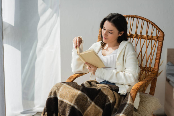 brunette woman in white cardigan reading novel in whicker chair under warm blanket - Fotografie, Obrázek
