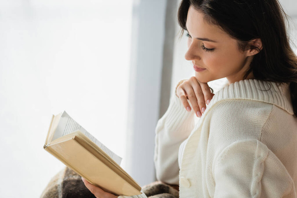 young woman in knitted cardigan reading novel at home - Фото, изображение