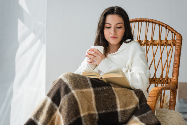 brunette woman sitting in wicker chair with cup of tea and reading novel - Fotoğraf, Görsel
