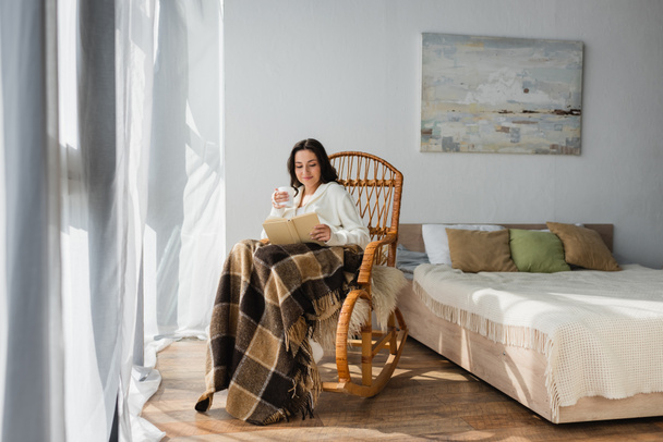 brunette woman reading book in bedroom while sitting in rocking chair under plaid blanket - Foto, Imagem