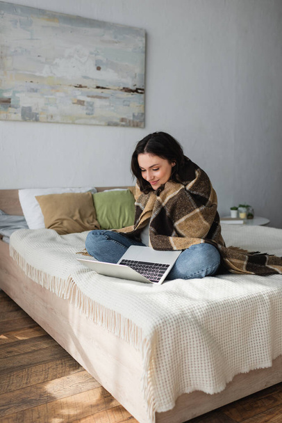 smiling woman sitting on bed under plaid blanket and looking at laptop  - Fotó, kép