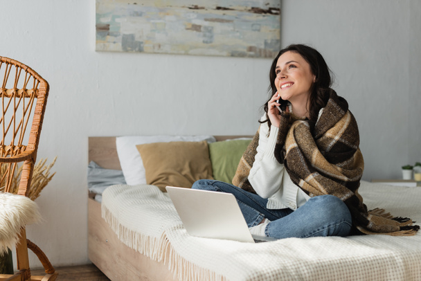 mujer alegre sentada en la cama cerca de la computadora portátil y hablando por teléfono móvil - Foto, Imagen