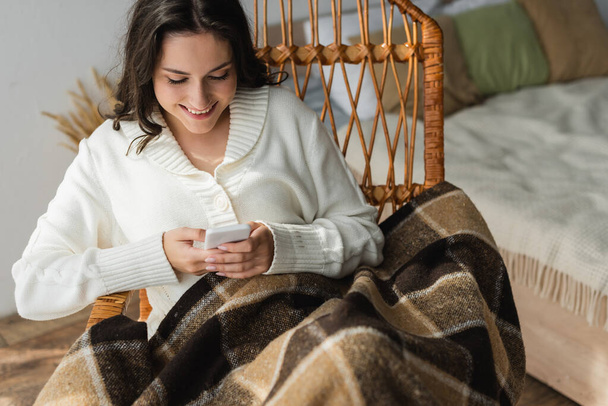 cheerful woman sitting in wicker chair under checkered blanket and typing on mobile phone - Photo, image