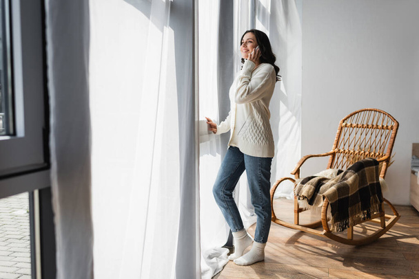 full length view of woman in warm cardigan standing near window and talking on cellphone - Photo, Image
