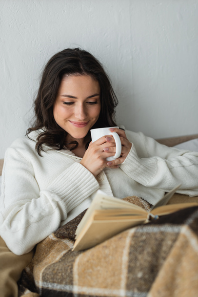 happy woman in warm cardigan drinking warm tea and reading book in bed - Фото, изображение