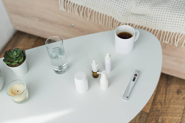 Ansicht von Behältern und Sprühflaschen mit Medikamenten in der Nähe von Getränken und Pflanzen auf dem Nachttisch - Foto, Bild