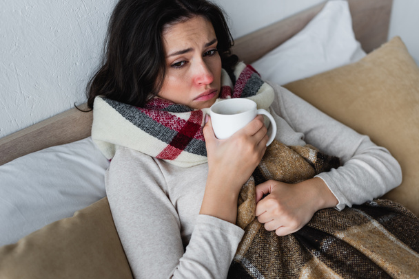 ill and upset woman lying under checkered blanket with cup of warm beverage - Foto, Imagem