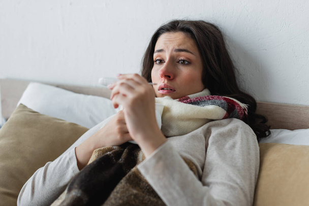 потрясенная и расстроенная женщина лежит под теплым одеялом и смотрит на термометр - Фото, изображение
