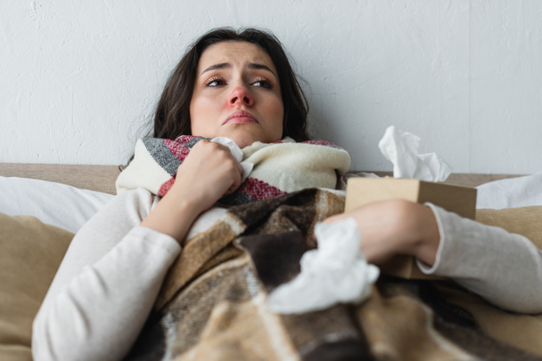 femme bouleversée couchée sous une couverture à carreaux près de serviettes en papier froissées, premier plan flou - Photo, image