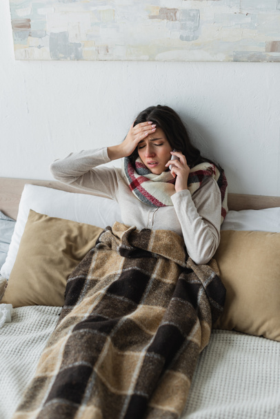 άρρωστη γυναίκα με κλειστά μάτια που μιλάει στο smartphone ενώ υποφέρει από πονοκέφαλο στο κρεβάτι - Φωτογραφία, εικόνα
