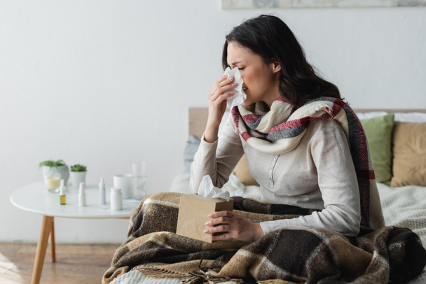mujer enferma en bufanda caliente sentada debajo de manta a cuadros y estornudos en servilleta de papel - Foto, imagen