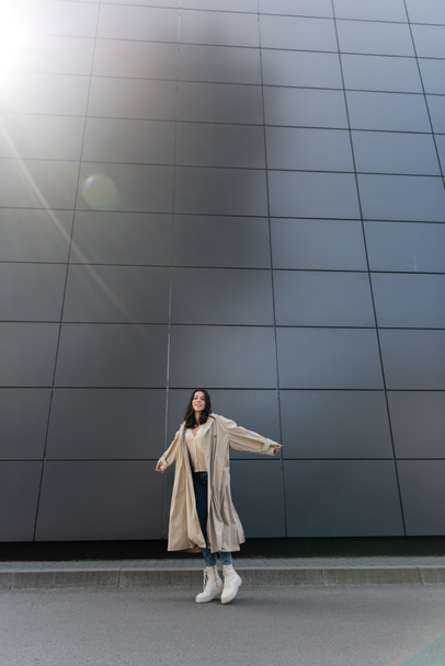 счастливая женщина в стильном осеннем наряде позирует с протянутыми руками возле высокой серой стены - Фото, изображение