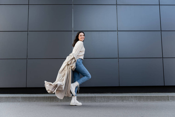mulher feliz em jeans e botas brancas sorrindo para a câmera enquanto corre ao longo da parede cinza - Foto, Imagem