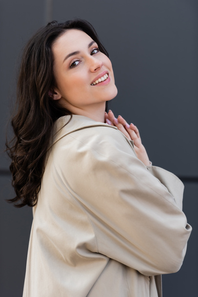 happy brunette woman in beige coat looking at camera outdoors - Photo, Image