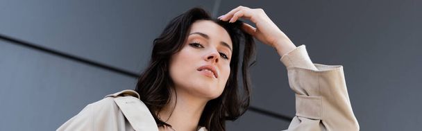 low angle view of brunette woman adjusting hair while looking at camera, banner - Фото, изображение