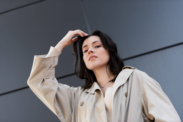 nízký úhel pohledu brunetky žena v béžovém kabátu pózuje s rukou v blízkosti hlavy venku - Fotografie, Obrázek