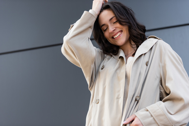 happy brunette woman in beige raincoat posing with closed eyes near grey wall - Фото, изображение