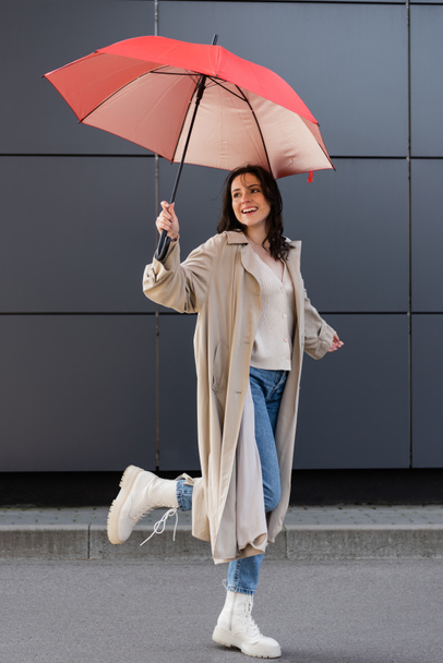 joyful woman in stylish autumn outfit standing on one leg under red umbrella outdoors - Photo, Image