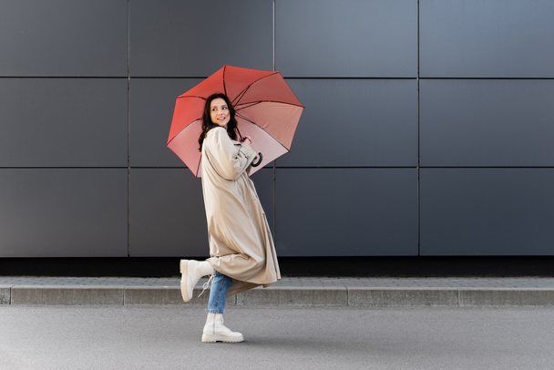 brunette vrouw in witte laarzen en beige jas terugkijkend onder rode paraplu - Foto, afbeelding