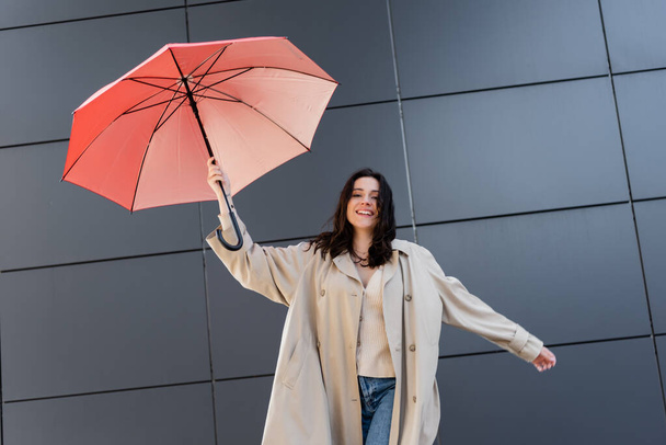 onnellinen brunette nainen beige takki katselee kameraa pitäen punainen sateenvarjo lähellä harmaa seinä - Valokuva, kuva