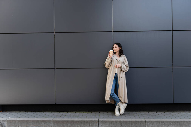 young woman in long raincoat standing with coffee to go near grey wall - Photo, image