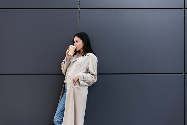 brunette woman drinking coffee while standing near grey wall with hand in pocket of beige coat - Foto, imagen